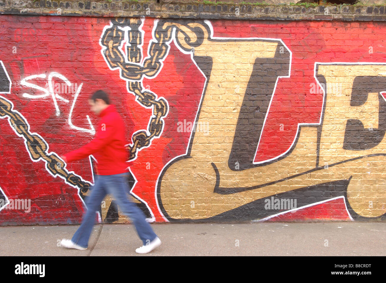 Un piéton promenades le long d'un trottoir à côté d'un graffitied wall dans Brick Lane, London. Banque D'Images