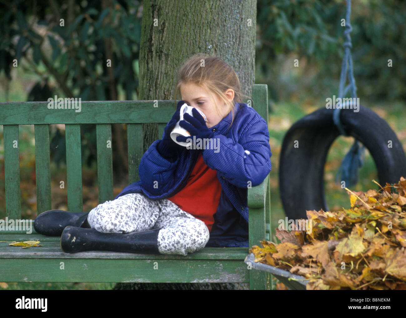 Petite fille assise dans le jardin réchauffement climatique les mains sur une boisson chaude après avoir recueilli une brouette pleine de feuilles Banque D'Images