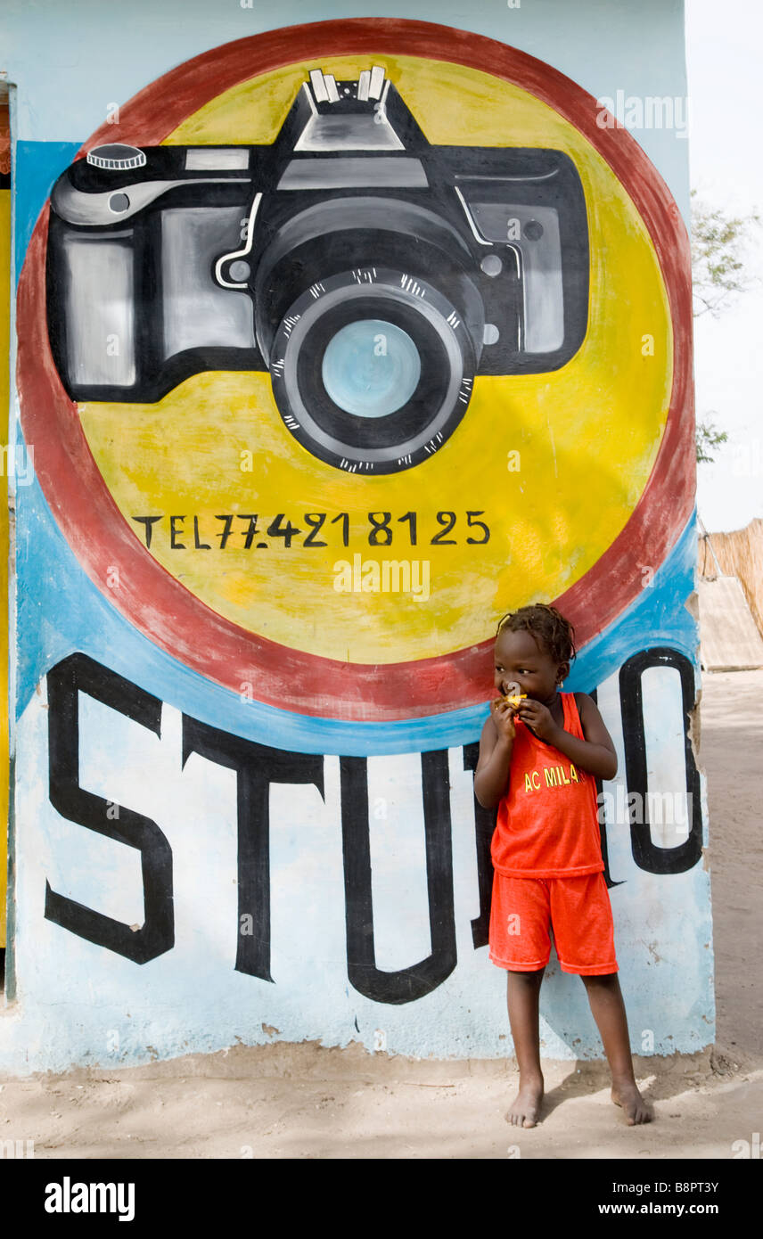 Petite fille de manger un morceau de mangue dans la rue de Toubacouta en face du studio photographique wall peintes au Sénégal Afrique de l'Ouest Banque D'Images