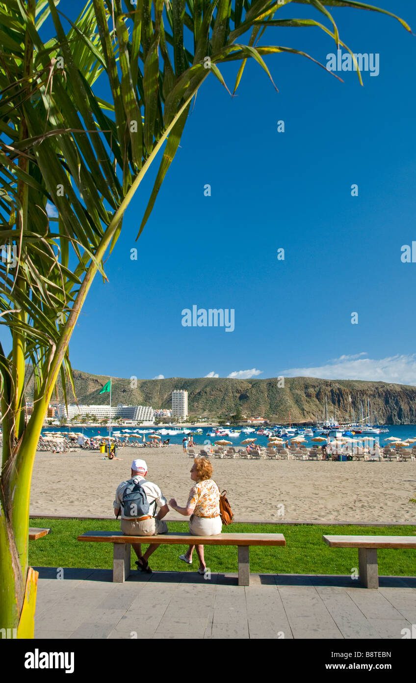 Couple de retraités âgés assis sur banc avec vue sur la plage de Los Cristianos encadrée par palmier typique sud de Tenerife Espagne Banque D'Images