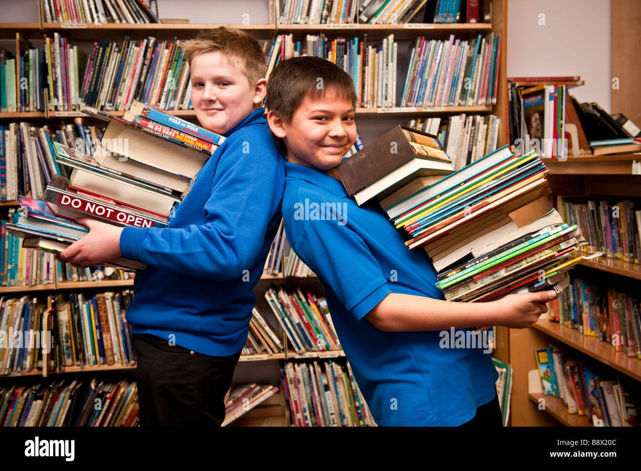 2 An 8 garçons dans la bibliothèque d'une école secondaire de Galles UK holding des piles de livres dans leurs bras Banque D'Images