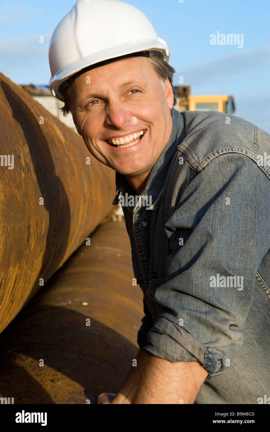Une belle quarantaine builder portant un casque est souriant comme il l'appuie contre certains gros tuyaux d'acier. Banque D'Images