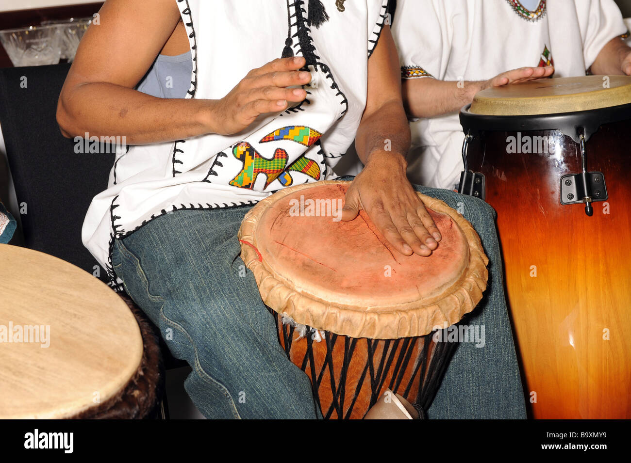 Homme jouant du tambour de la peau animale africaine avec un autre homme jouant un grand tambour Congo tant en manche courte chemise blanche et jeans Banque D'Images