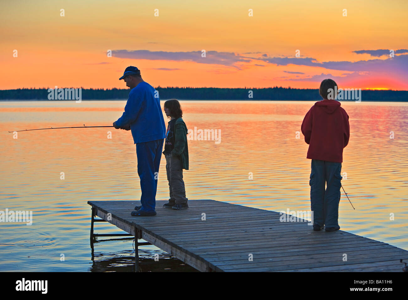 Un père avec ses deux fils de pêche à la fin d'un quai sur le lac Audy au coucher du soleil,le parc national de Riding Mountain, Manitoba,Canada. Banque D'Images