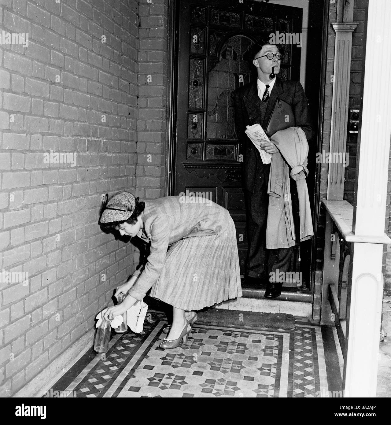 1950s, un couple quitte la maison, l'homme avec pipe et imperméable, la dame mettant des bouteilles de lait vides dans le porche couvert pour la collecte, Angleterre, Royaume-Uni. Banque D'Images