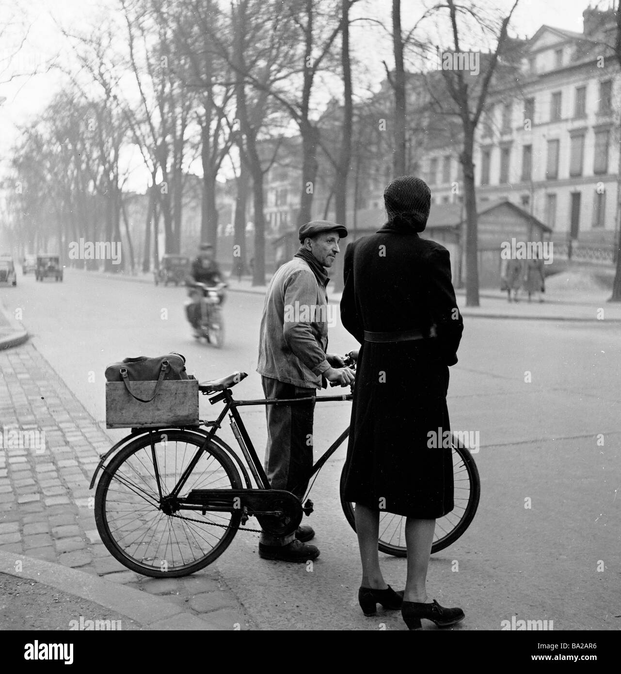 Paris, France, 1950s. Une femme de grande taille portant un manteau debout  à côté d'un homme portant une casquette plate et avec son vélo attendant de  traverser une route Photo Stock -