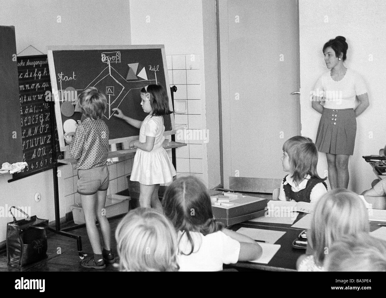 Années 70, photo en noir et blanc, l'édification, l'école, des écoliers et des écolières dans une classe d'école pendant les cours, l'enseignante, les enfants âgés de 7 à 10 ans, garçon et fille à un tableau noir Banque D'Images