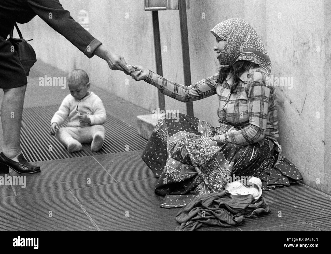 Années 70, photo en noir et blanc, personnage, pauvreté, mendiant-femme est assise au bord de la route, son bébé joue sur une grille, d'une femme présente ses gamins, âgés de 25 à 30 ans, de 6 à 12 mois, l'Italie, Lombardie, Milan Banque D'Images