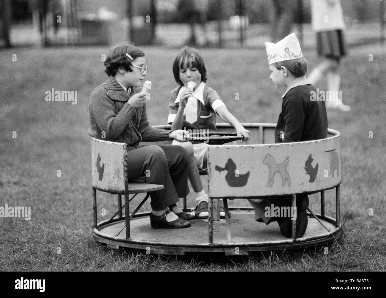 Années 70, photo en noir et blanc, les gens, les enfants, les traiter pour enfants, deux filles et un garçon avec un chapeau de papier sur la tête s'asseoir dans un rond-point et lécher la crème glacée, de 5 à 8 ans Banque D'Images