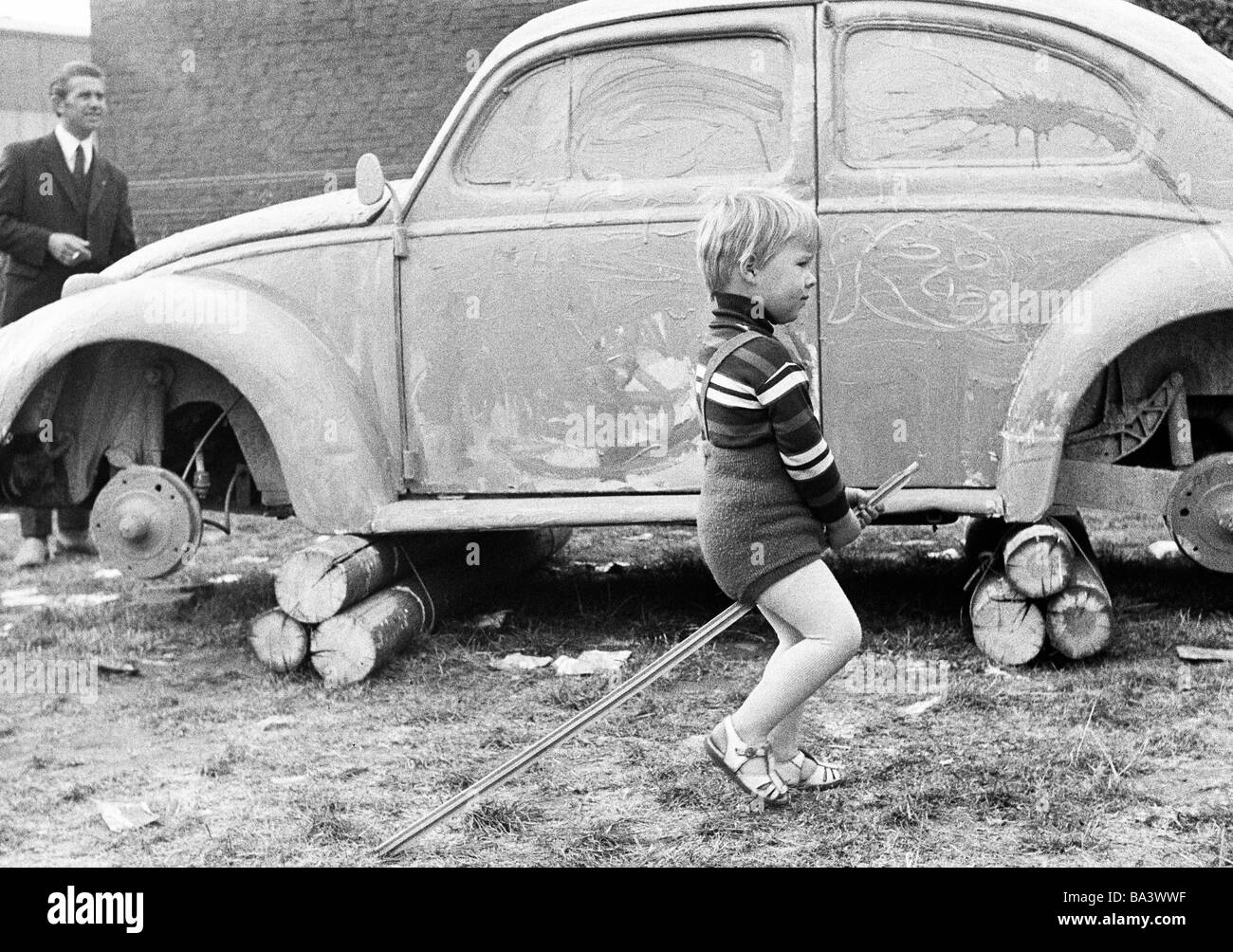 Années 70, photo en noir et blanc, les gens, les enfants, petit garçon se déplace sur un bâton, de jeu, de 3 à 4 ans, dans l'arrière-plan d'une voiture avec un cric et peint sans roues, VW Coccinelle Banque D'Images