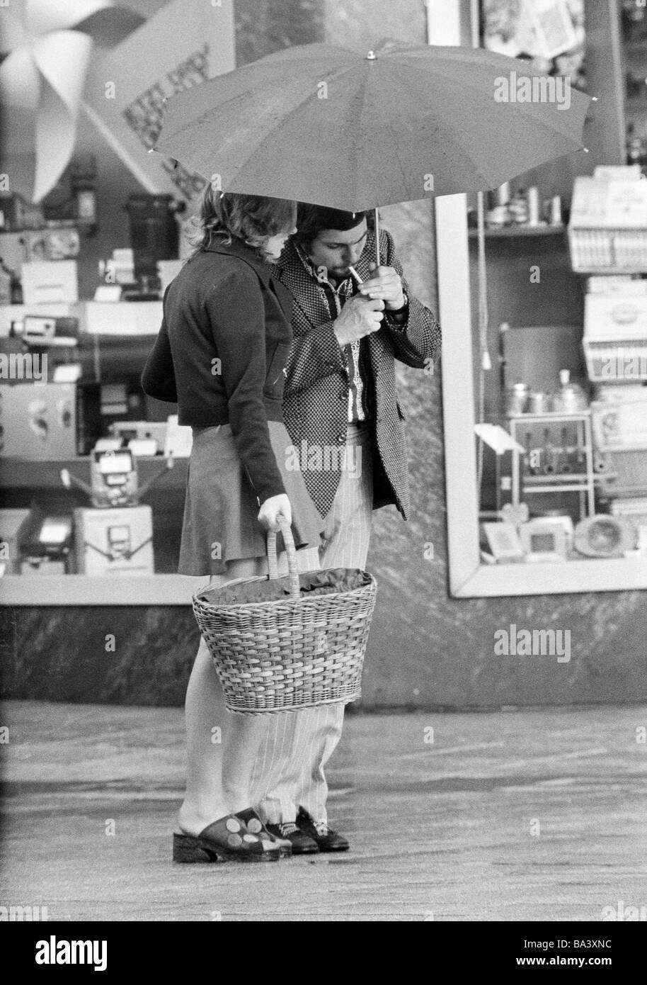 Années 70, photo en noir et blanc, météo, pluie, jeune couple bras dessus bras dessous sous un parapluie, l'homme s'allume une cigarette, âgés de 20 à 30 ans Banque D'Images