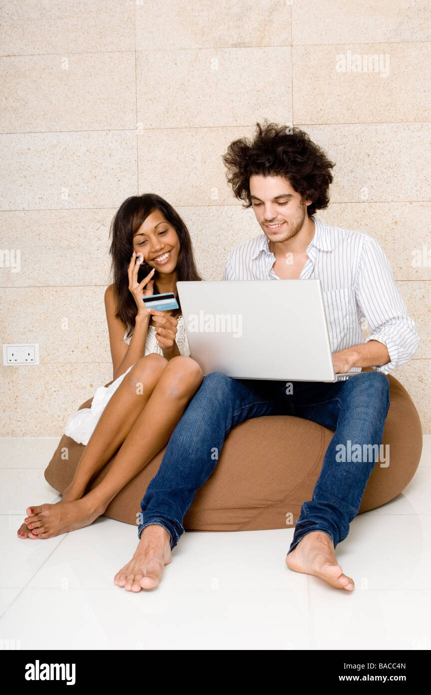 Jeune homme assis sur un sac de haricots à l'aide d'un ordinateur portable avec une jeune femme assise à côté de lui parler sur un téléphone mobile Banque D'Images