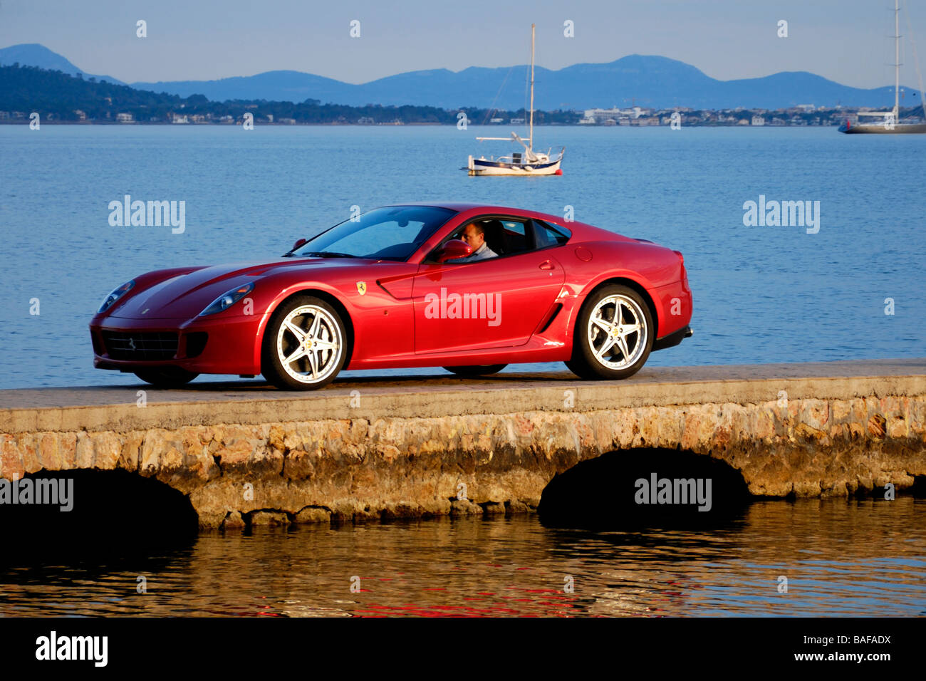 Rouge Ferrari avec chauffeur privé sur le dock de la Méditerranée dans la baie de Pollensa avec les yachts et les montagnes en arrière-plan au coucher du soleil Banque D'Images