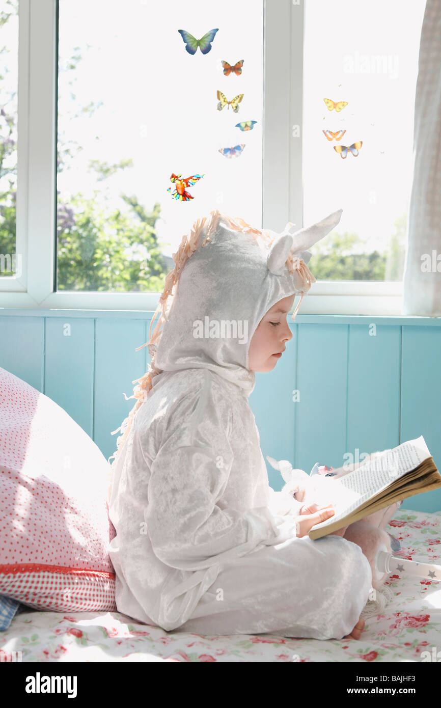 Jeune fille (5-6) sitting on bed in costume unicorn reading book, side view Banque D'Images
