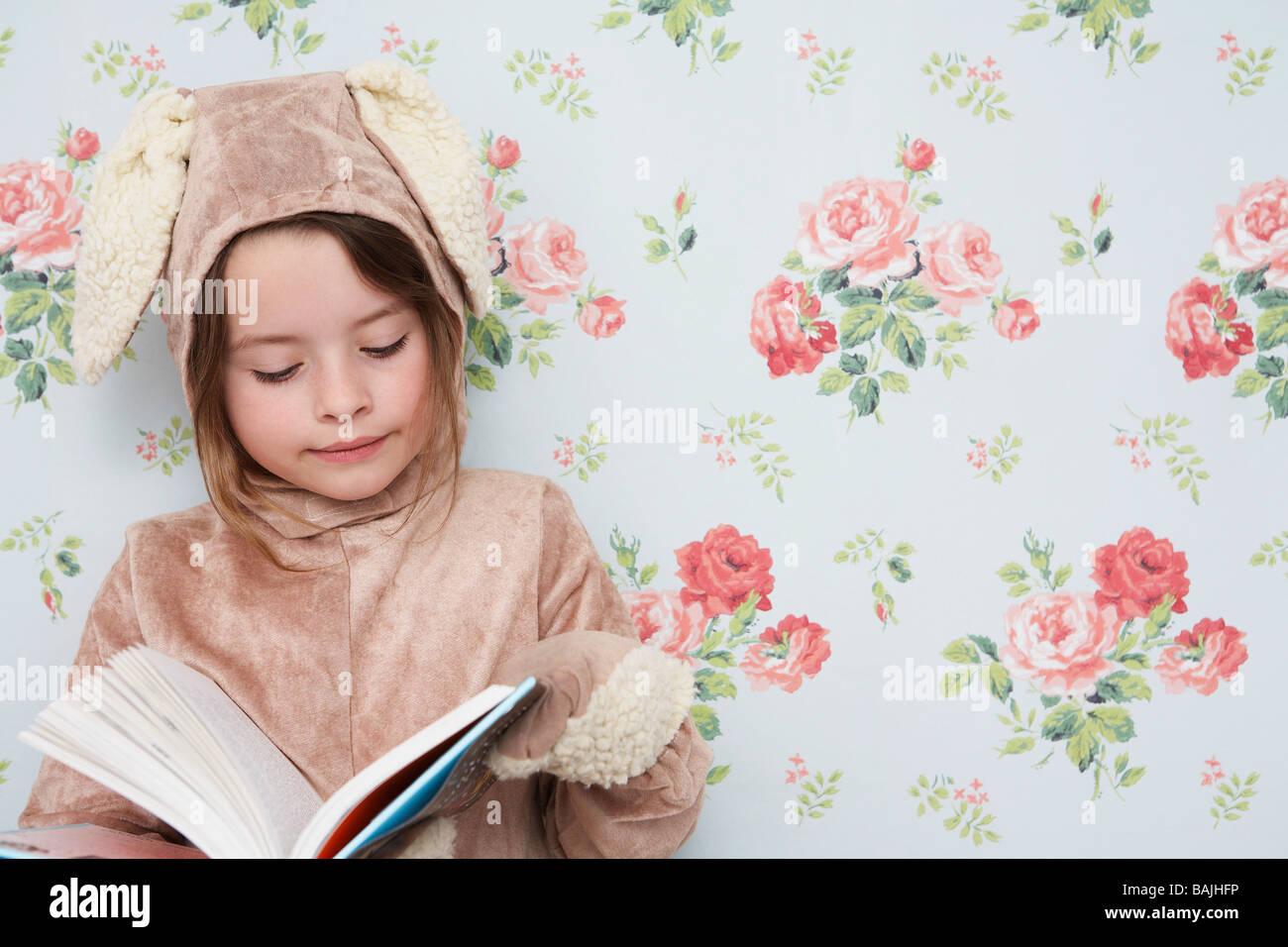 Jeune fille (5-6) n costume de lapin reading book, papier peint Motif floral avec en arrière-plan Banque D'Images