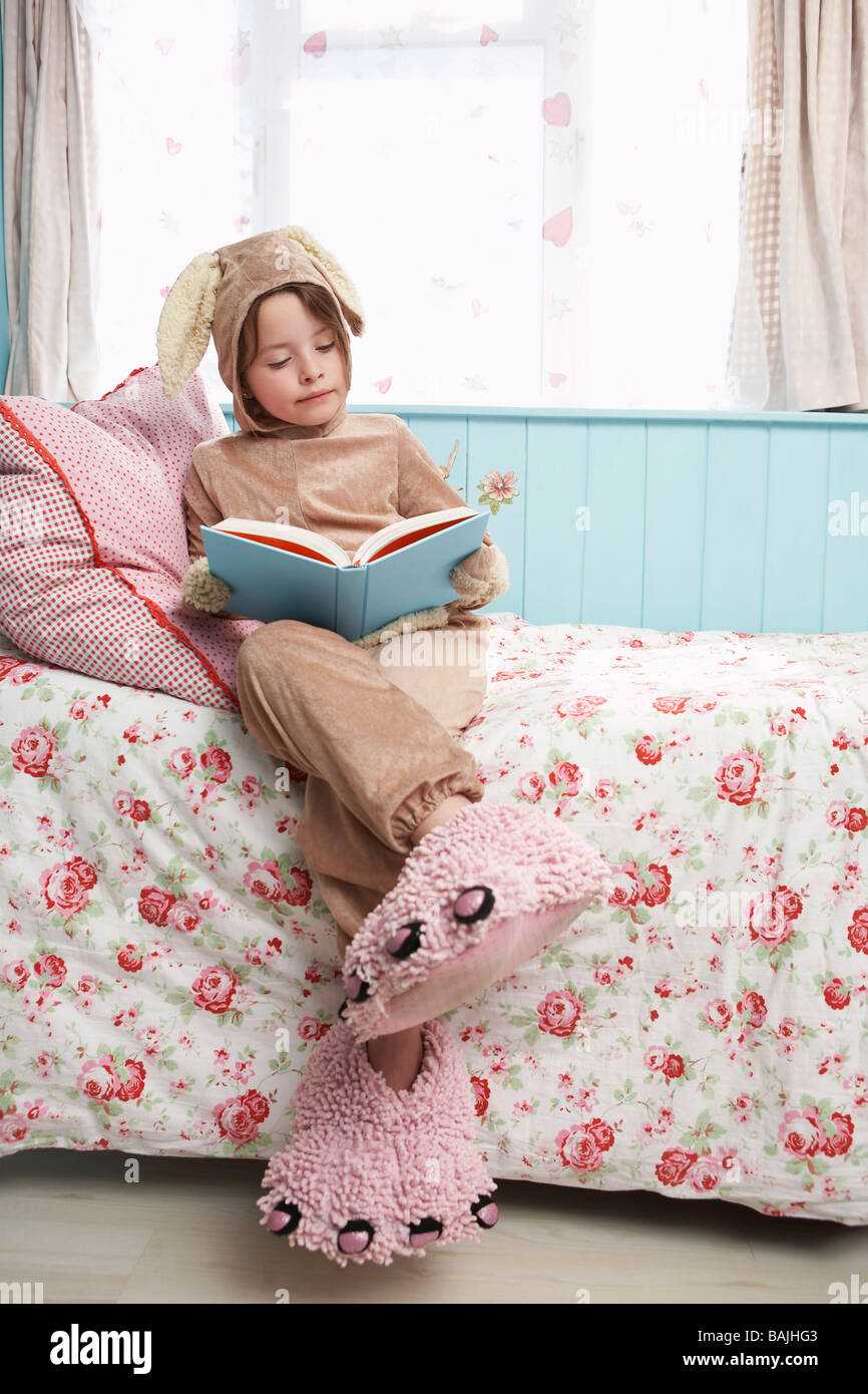 Jeune fille (5-6) sitting on bed wearing costume de lapin et monster chaussons, reading book Banque D'Images