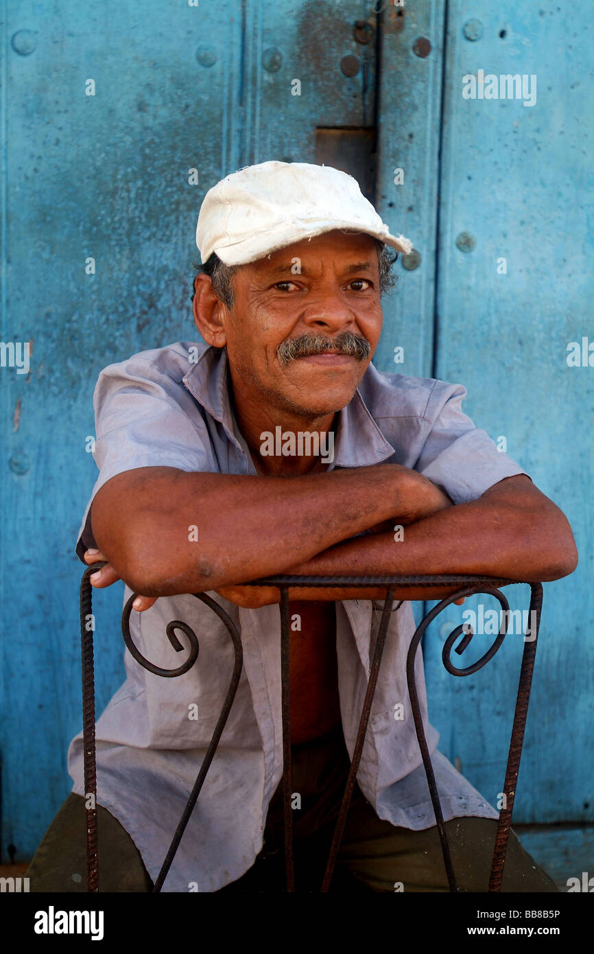 Un portrait d'un homme portant un senior cubaine casquette blanche  s'appuyant sur l'arrière du dossier de chaise en fer forgé peint bleu par  porte. Trinidad, Cuba, Caraïbes Photo Stock - Alamy