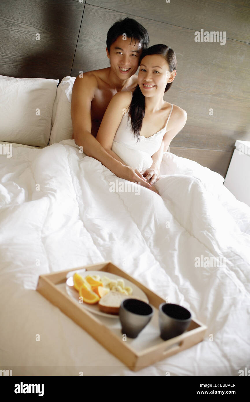 Couple assis dans son lit, looking at camera, plateau du petit déjeuner au pied du lit Banque D'Images
