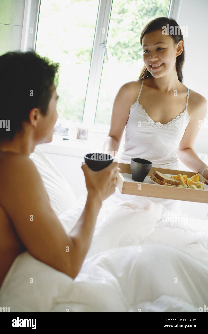 Woman holding plateau du petit déjeuner, l'homme assis au lit holding cup Banque D'Images