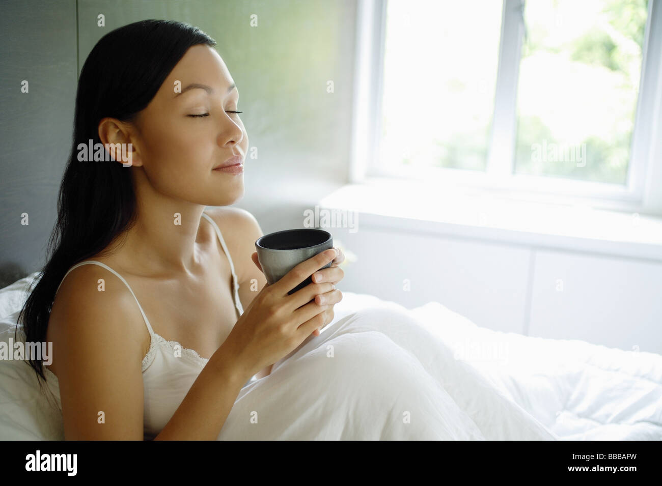 Femme assise dans son lit, les yeux fermés, holding cup Banque D'Images