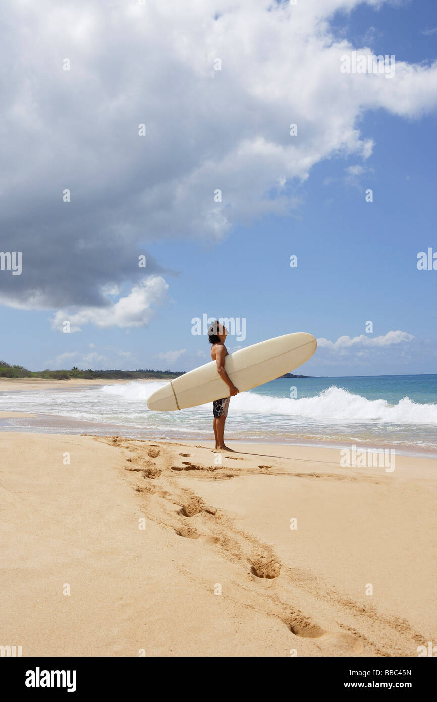 Asian man standing on beach, à Hawaii holding surfboard, à la recherche de sky Banque D'Images