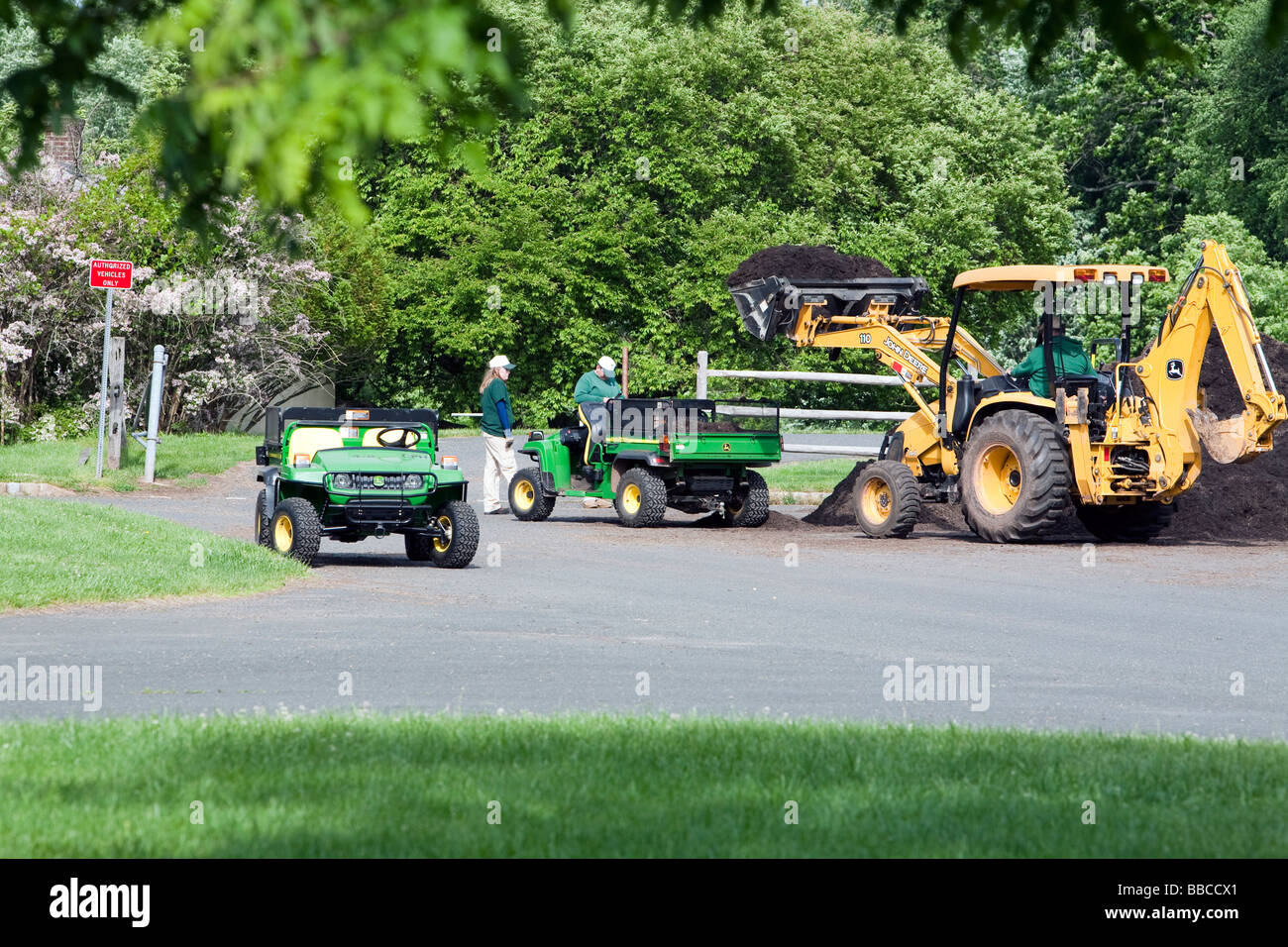 Jardiniers travaillant avec et pour l'épandage de paillis de chargement. Un tracteur John Deere jaune et varous les travailleurs et les charrettes et les VTT. Banque D'Images