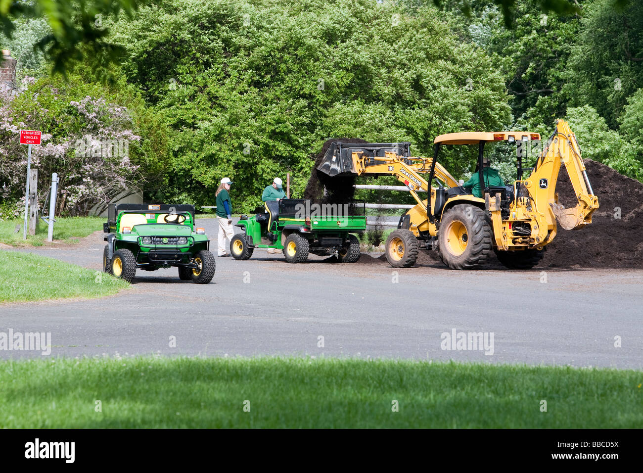 Jardiniers travaillant avec et pour l'épandage de paillis de chargement. Un tracteur John Deere jaune et varous les travailleurs et les charrettes et les VTT. Banque D'Images