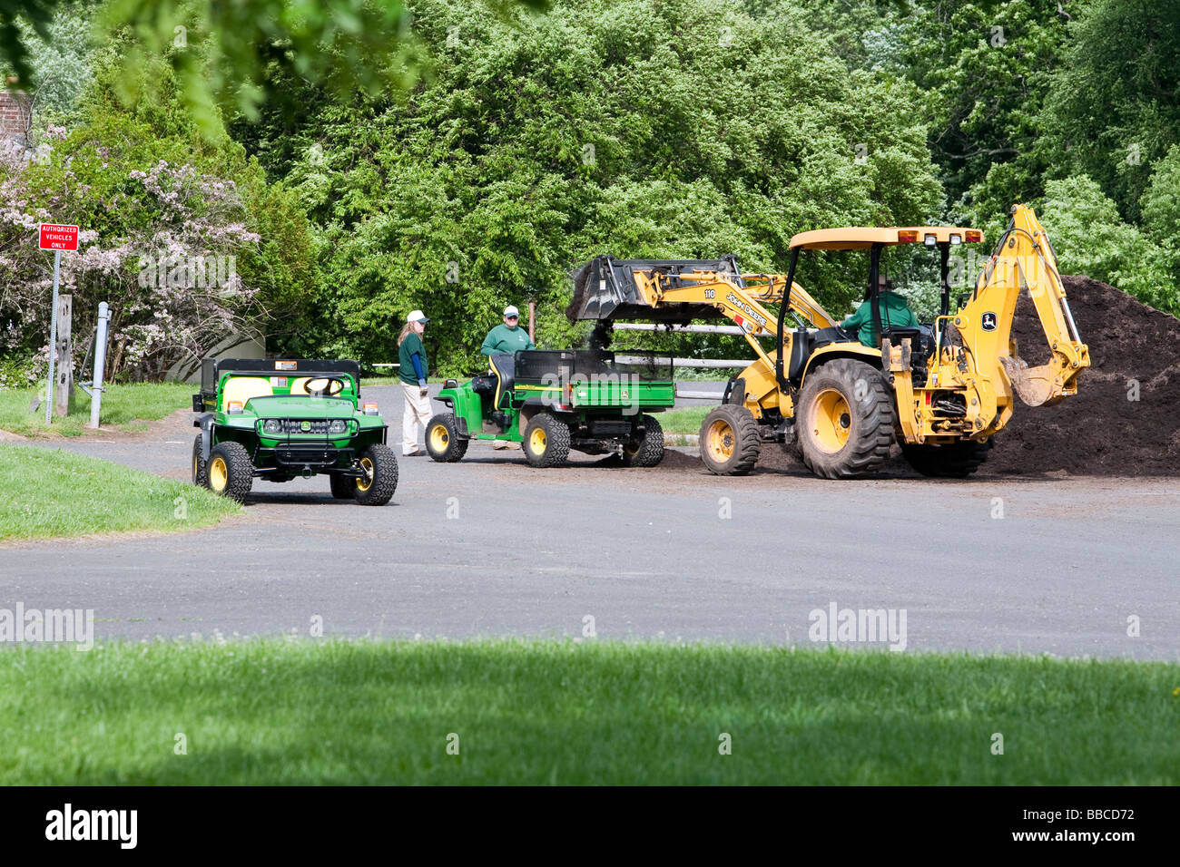 Jardiniers travaillant avec et pour l'épandage de paillis de chargement. Un tracteur John Deere jaune et varous les travailleurs et les charrettes et les VTT. Banque D'Images