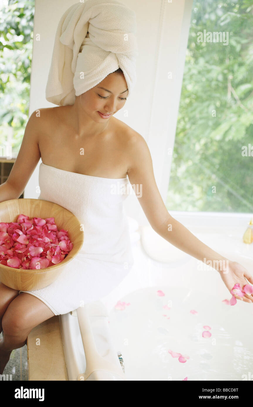 Femme en serviette, assis à bord de baignoire à remous, jetant des pétales de fleurs dans l'eau Banque D'Images