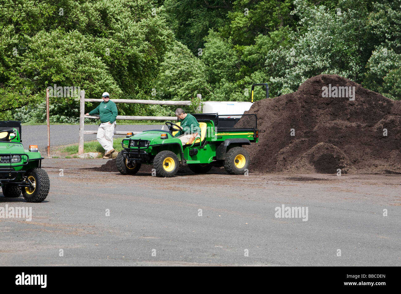 Jardiniers travaillant avec et pour l'épandage de paillis de chargement. Un tracteur John Deere jaune et varous les travailleurs et les charrettes et les VTT. Banque D'Images