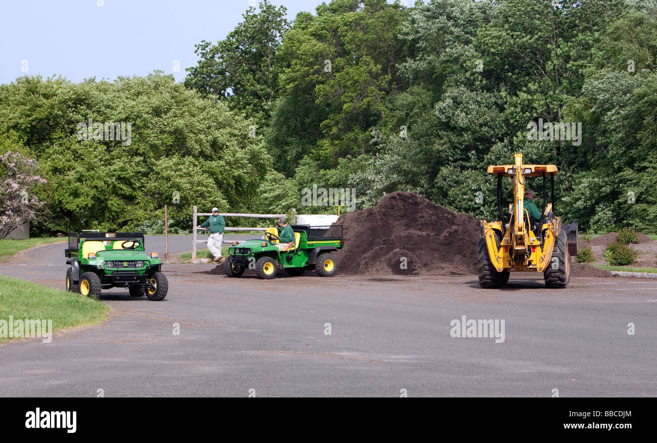 Jardiniers travaillant avec et pour l'épandage de paillis de chargement. Un tracteur John Deere jaune et varous les travailleurs et les charrettes et les VTT. Banque D'Images