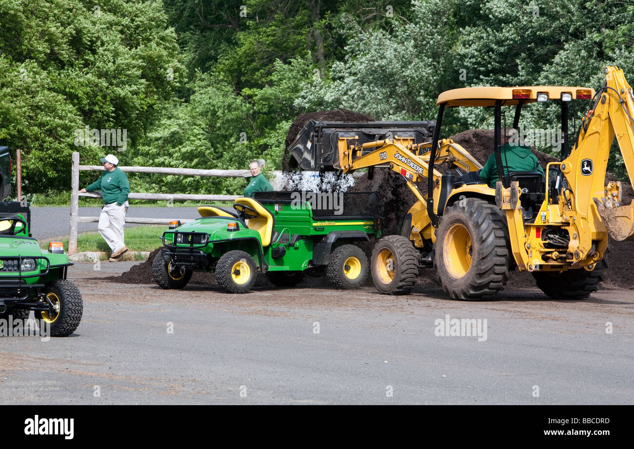 Jardiniers travaillant avec et pour l'épandage de paillis de chargement. Un tracteur John Deere jaune et varous les travailleurs et les charrettes et les VTT. Banque D'Images
