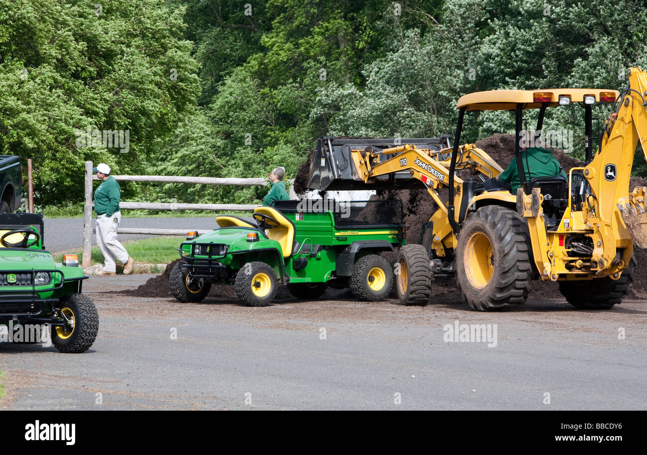 Jardiniers travaillant avec et pour l'épandage de paillis de chargement. Un tracteur John Deere jaune et varous les travailleurs et les charrettes et les VTT. Banque D'Images