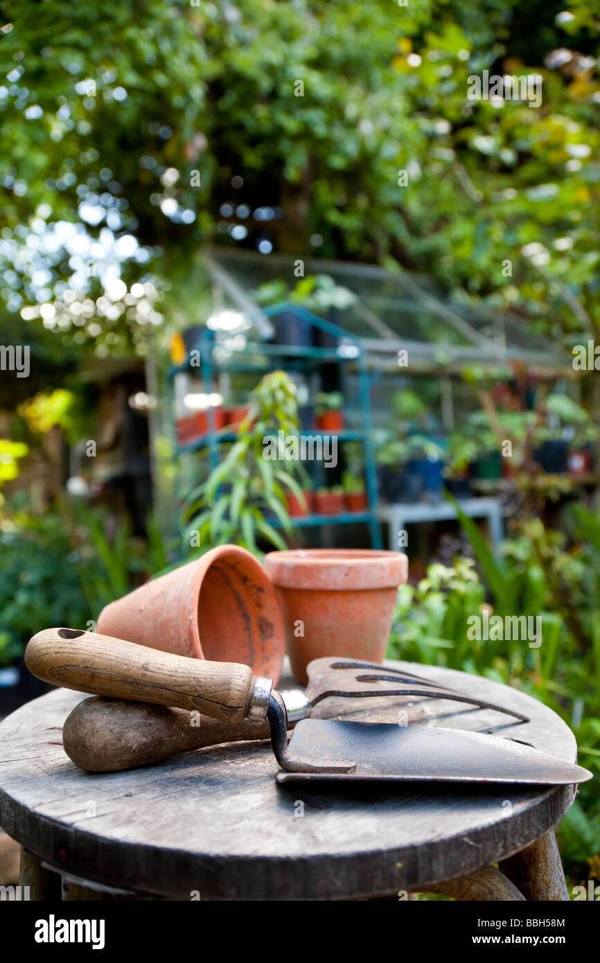 Outils de jardinage et les pots de fleurs reposant sur un tabouret dans un jardin verdoyant avec une serre non mise au point à l'arrière-plan Banque D'Images