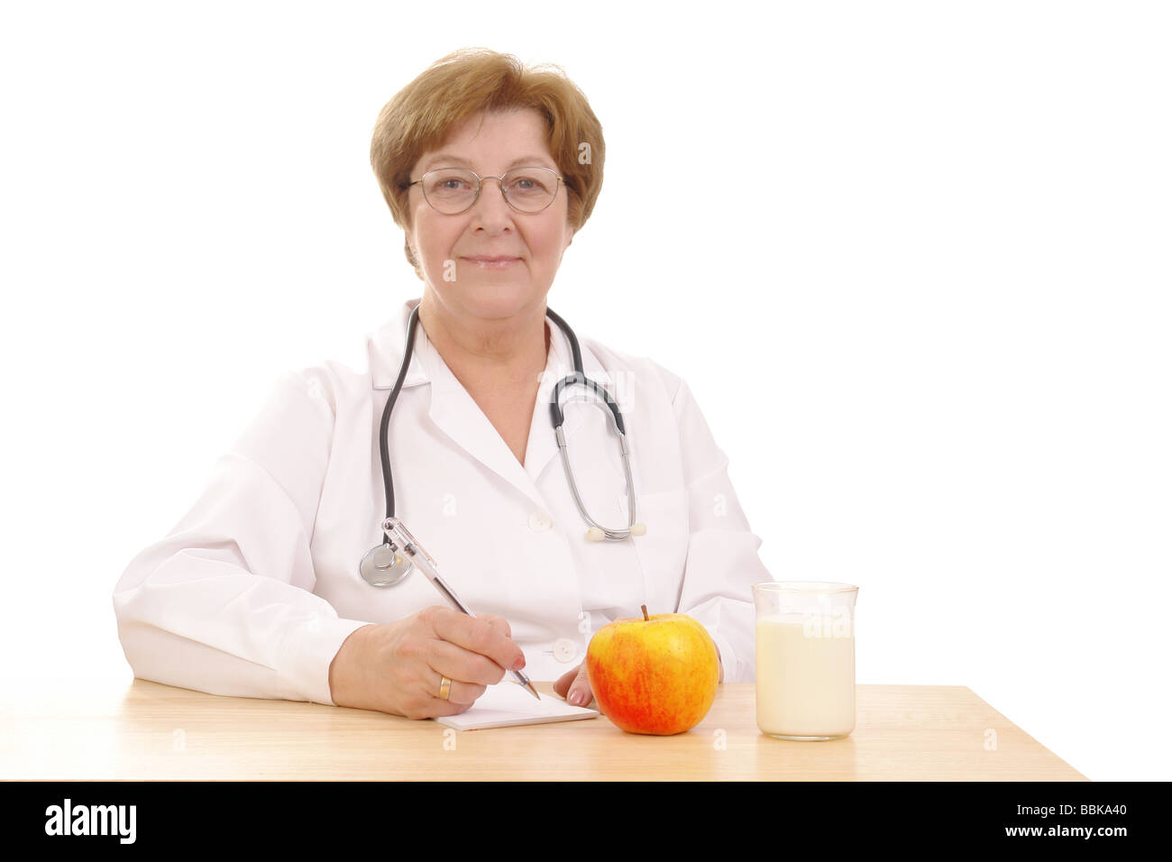 Senior female doctor sitting behind desk avec verre de lait et apple isolé sur fond blanc Banque D'Images