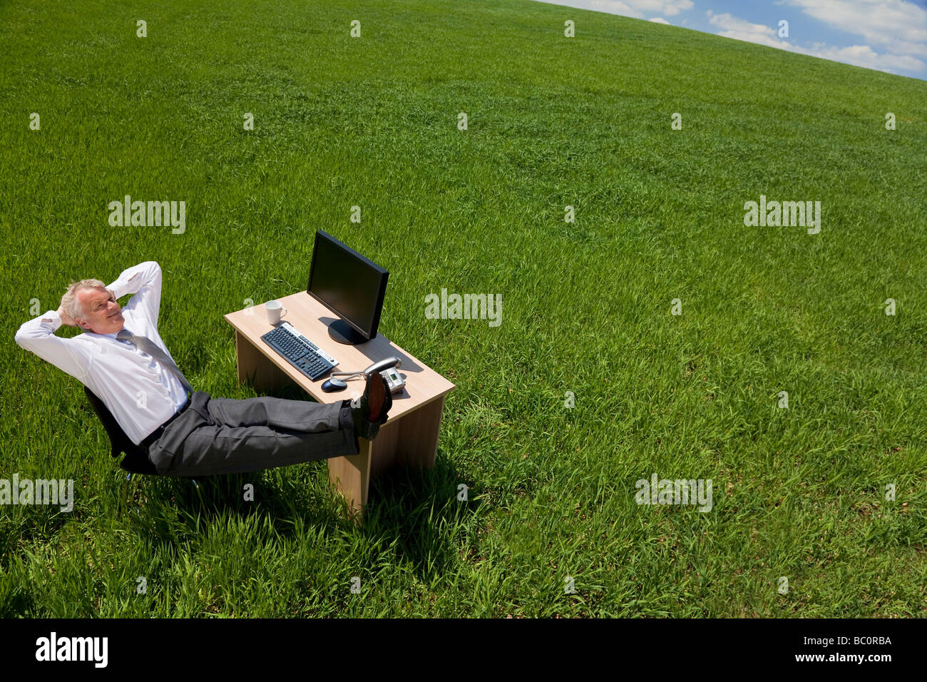 Concept d'affaires shot montrant un homme plus âgé de la détente à son bureau dans un champ vert avec un ciel bleu Banque D'Images