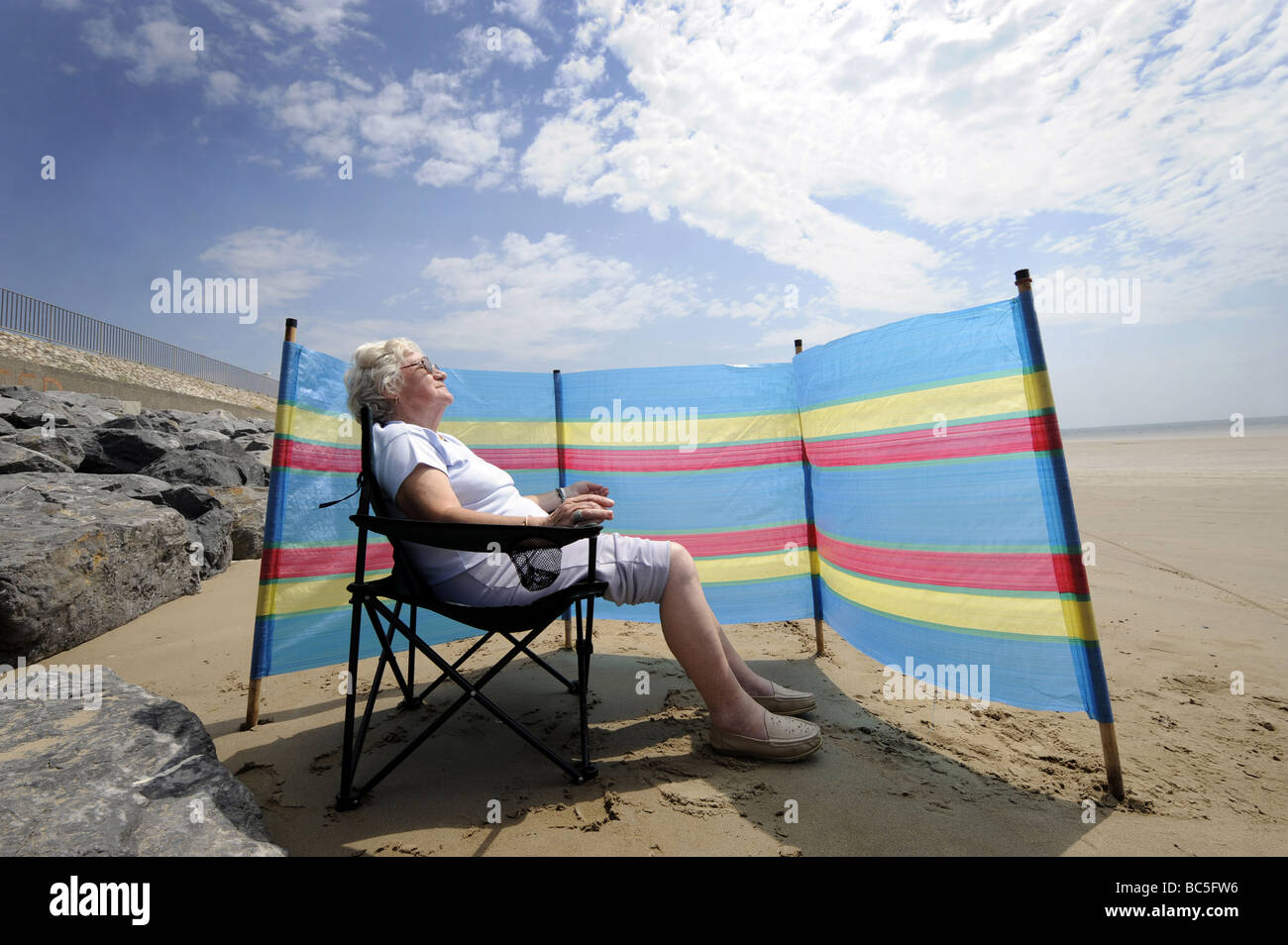 Un PENSIONNÉ BÉNÉFICIE D'un bain de soleil SUR UNE PLAGE BRITANNIQUE AVEC UN COUPE-VENT RE RETRAITE RETRAITÉS VACANCES SAVONS SUN DANGER UK Banque D'Images