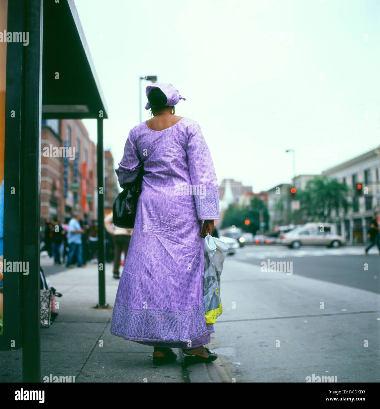 Vue arrière de la femme africaine sur textiles en attente à un arrêt de bus à Harlem en portant une robe violette achetés en Afrique, New York City USA KATHY DEWITT Banque D'Images