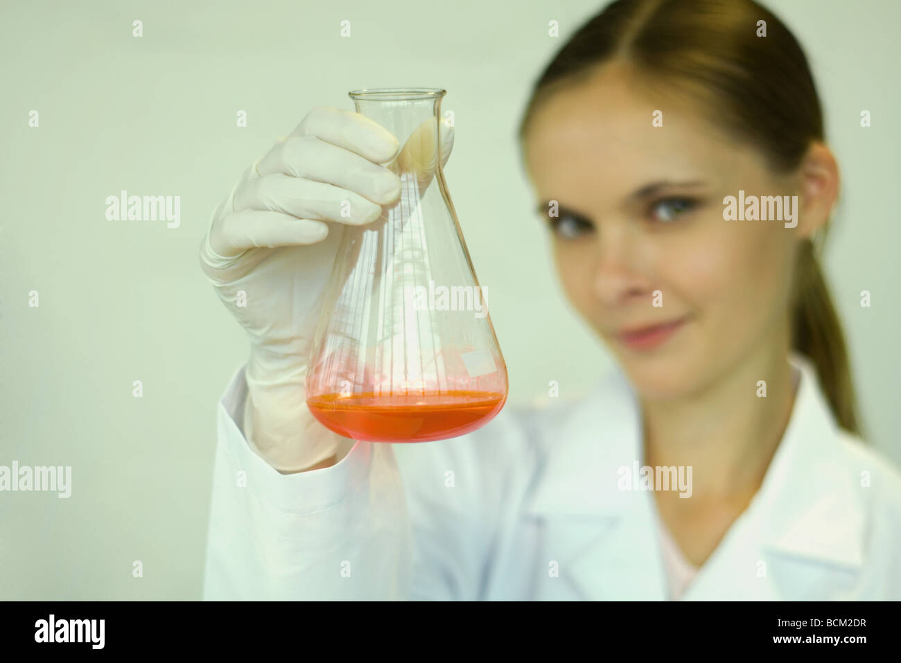Female scientist holding up bécher avec liquide à l'intérieur, looking at camera, focus on foreground Banque D'Images