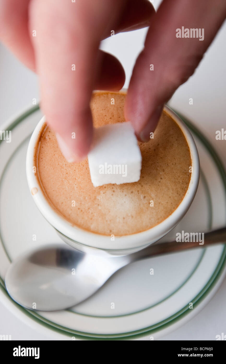 Vue aérienne d'une main féminine droping un cube de sucre dans la mousse d'un café expresso chaud boire dans une tasse avec une cuillère en métal Banque D'Images