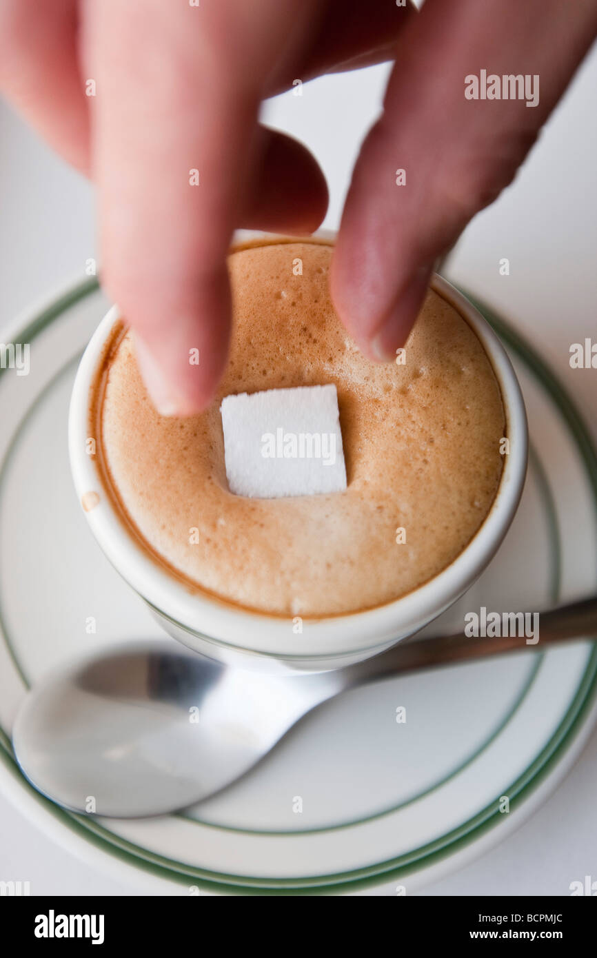 Vue aérienne d'une main féminine droping un cube de sucre dans la mousse d'un café expresso chaud boire dans une tasse avec une cuillère en métal Banque D'Images