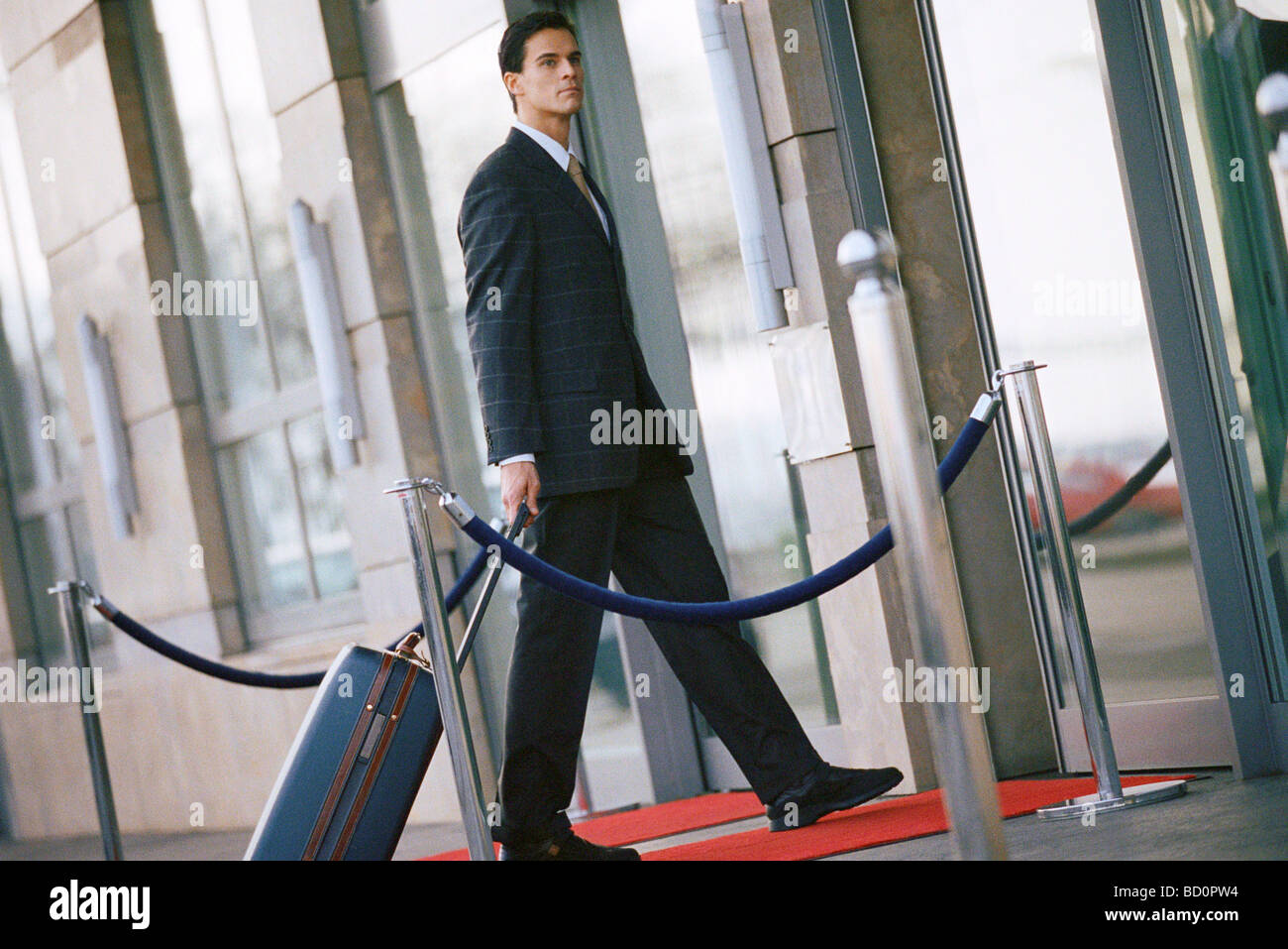 Businessman walking vers l'entrée du bâtiment, pulling suitcase derrière lui Banque D'Images