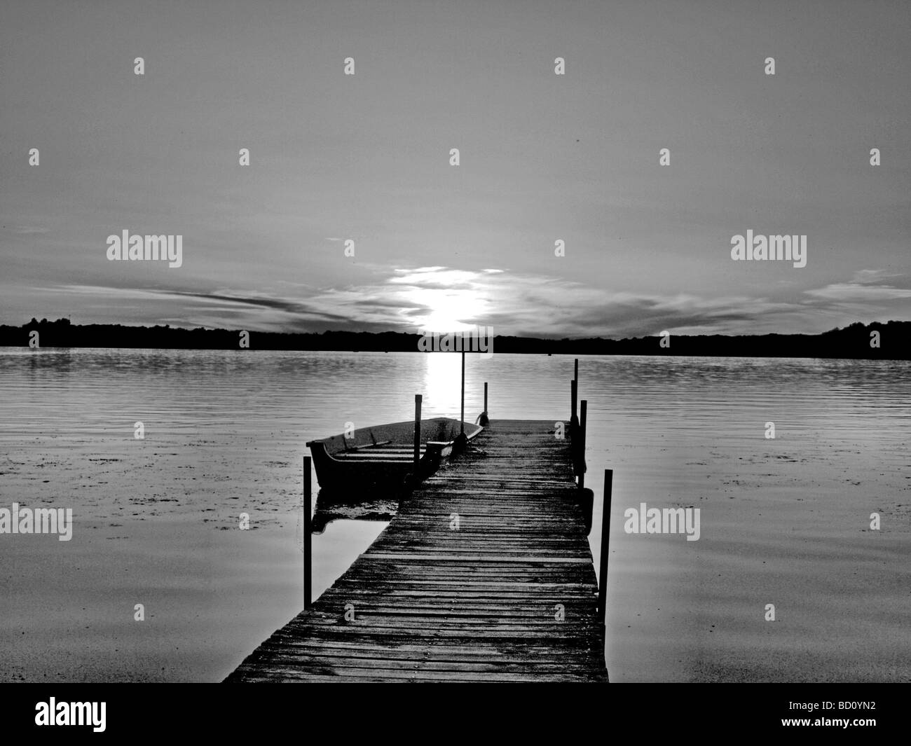 Vue de la rive sur le lac comme si on se tient à la fin d'un dock pier avec barque voile à la recherche sur un coucher de soleil et sur les rives, b & w Banque D'Images