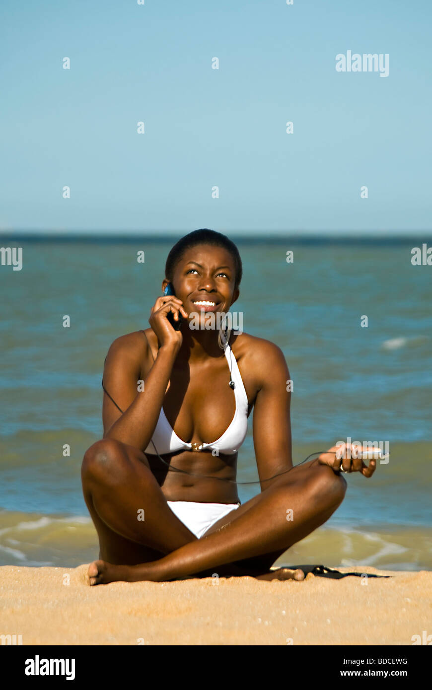 Portrait d'une jolie verticale femme africaine dans un bikini blanc sitting  cross legged en utilisant un téléphone mobile à la plage Photo Stock - Alamy