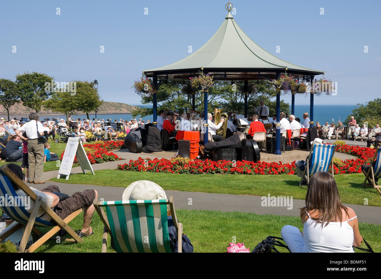 Vacanciers touristes assis dans des chaises longues à l'écoute d'un groupe de cuivres jouant dans le kiosque d'été Crescent Gardens Filey North Yorkshire Angleterre GB Banque D'Images