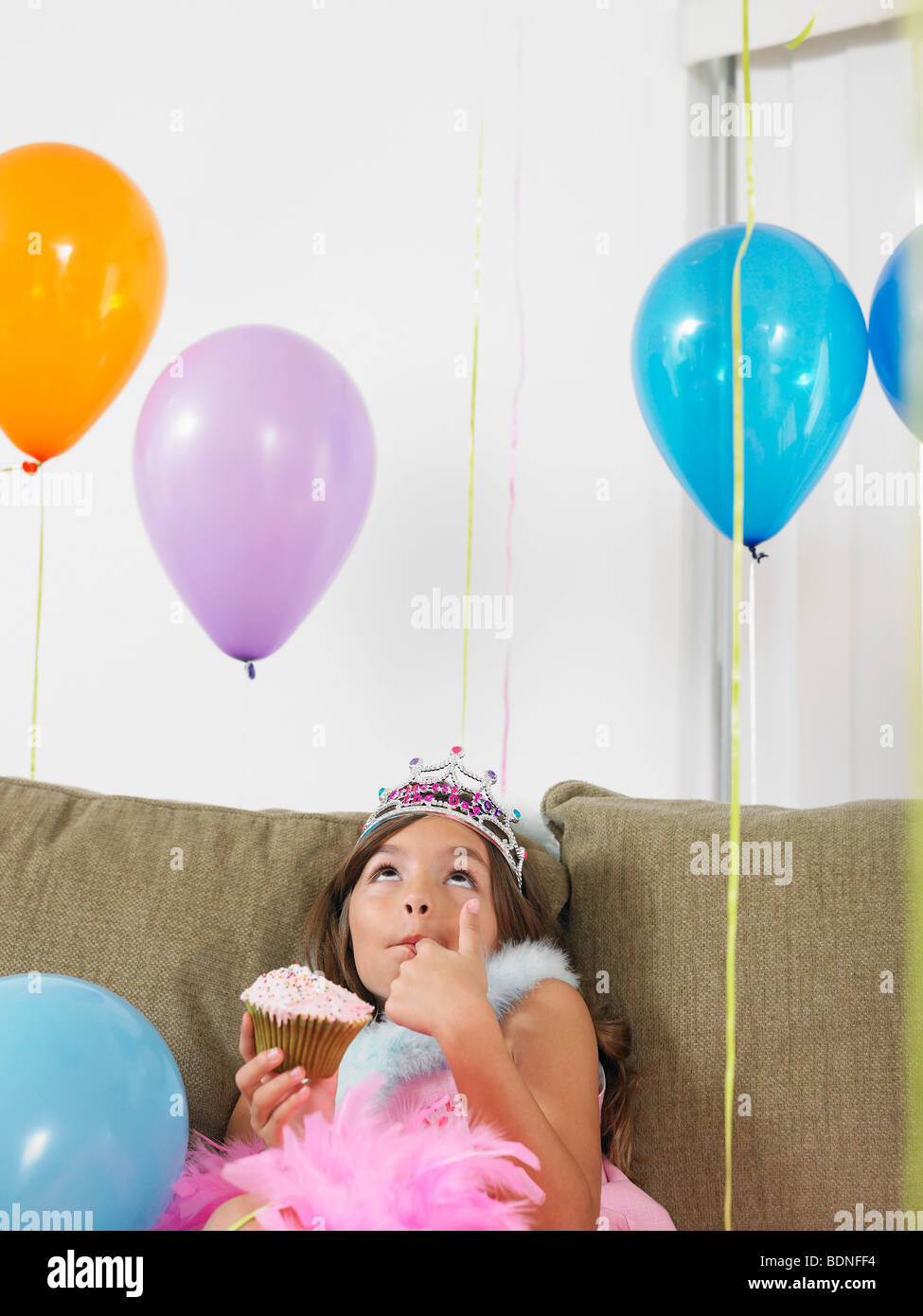 Jeune fille (7-9) sitting on sofa eating cupcake, looking up at balloons Banque D'Images