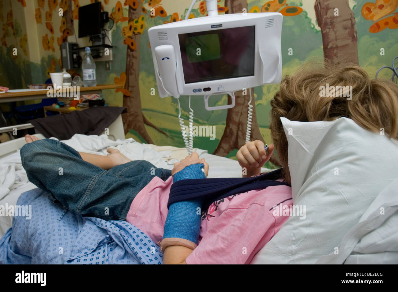 Un garçon avec un bras cassé se trouve sur un lit en regardant la télé en attente de traitement dans un hôpital du NHS Banque D'Images