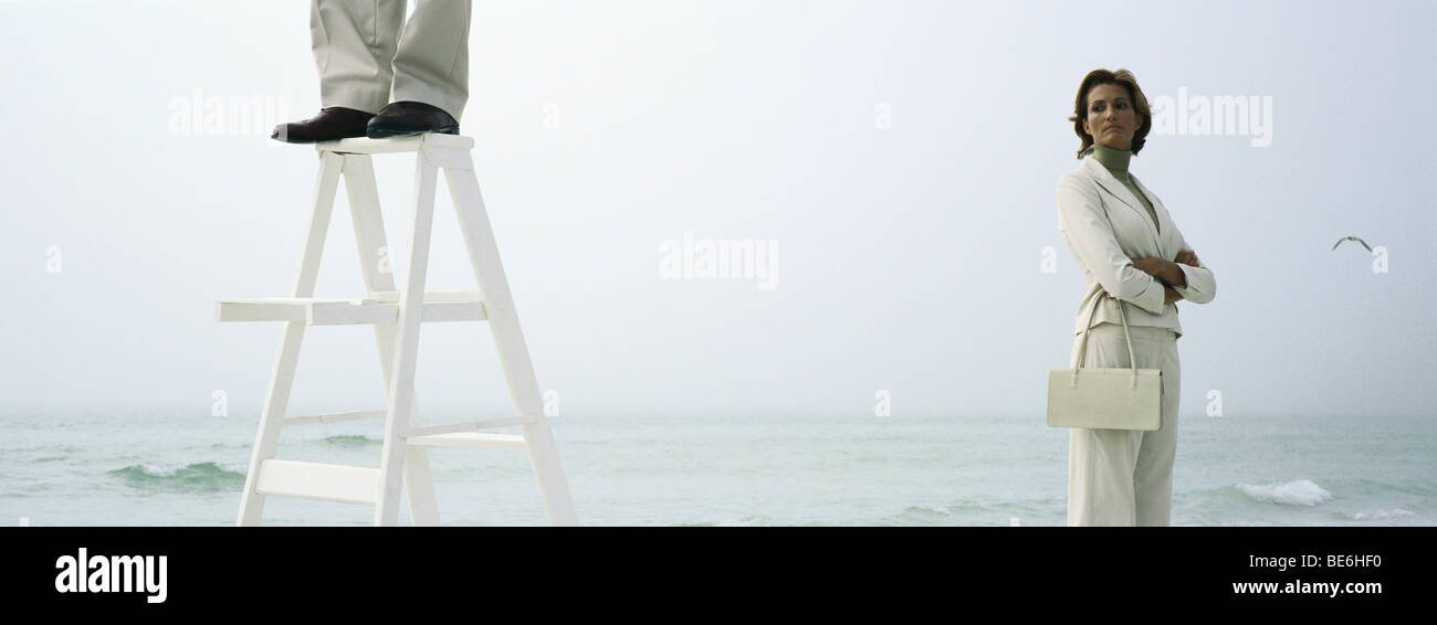 Woman on beach with folded arms watching homme debout sur le dessus de l'échelle, cropped Banque D'Images