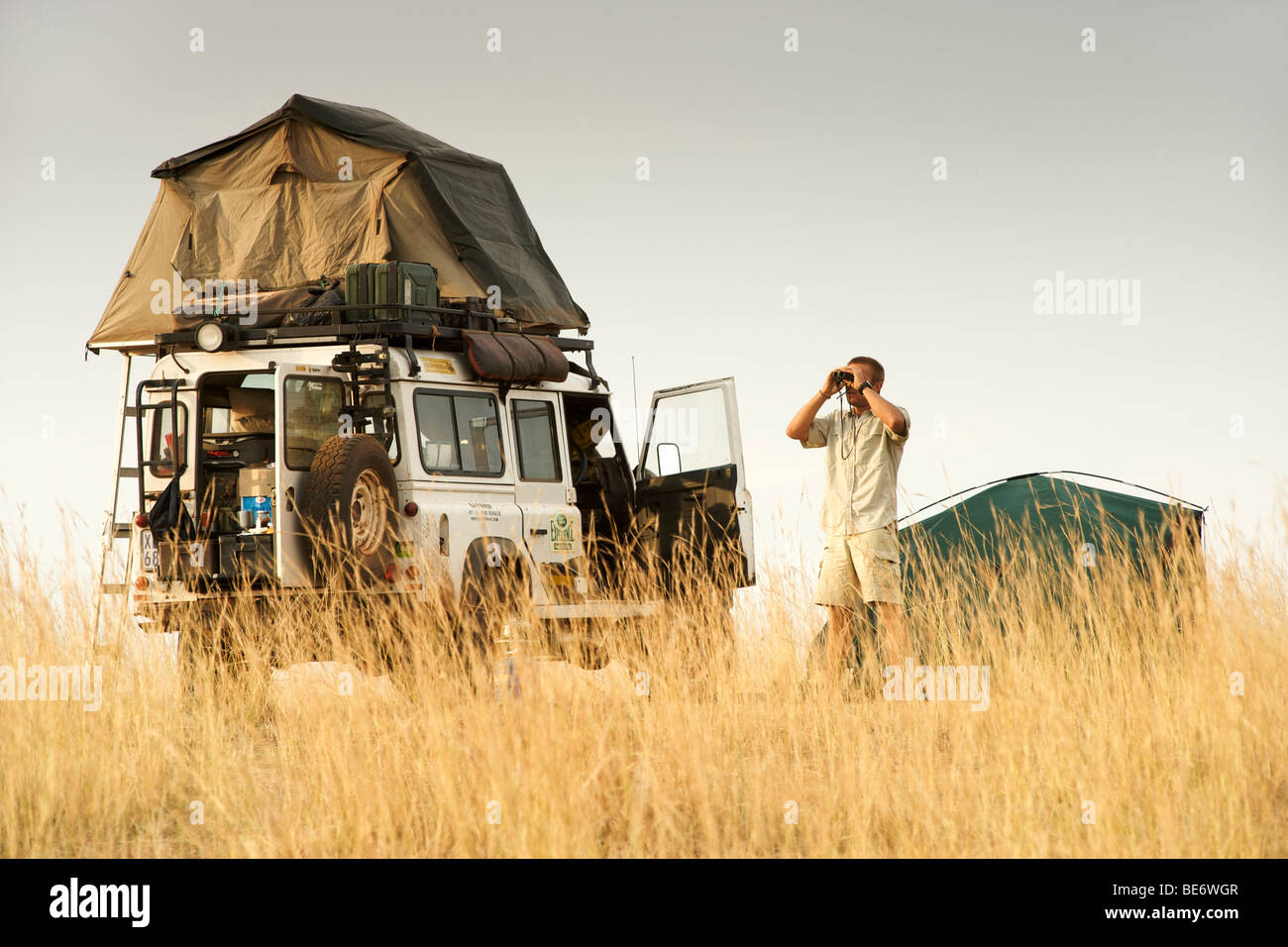Un homme regardant à travers des jumelles aux côtés d'un Land Rover Defender dans un camping dans la réserve faunique Kabwoya en Ouganda. Banque D'Images
