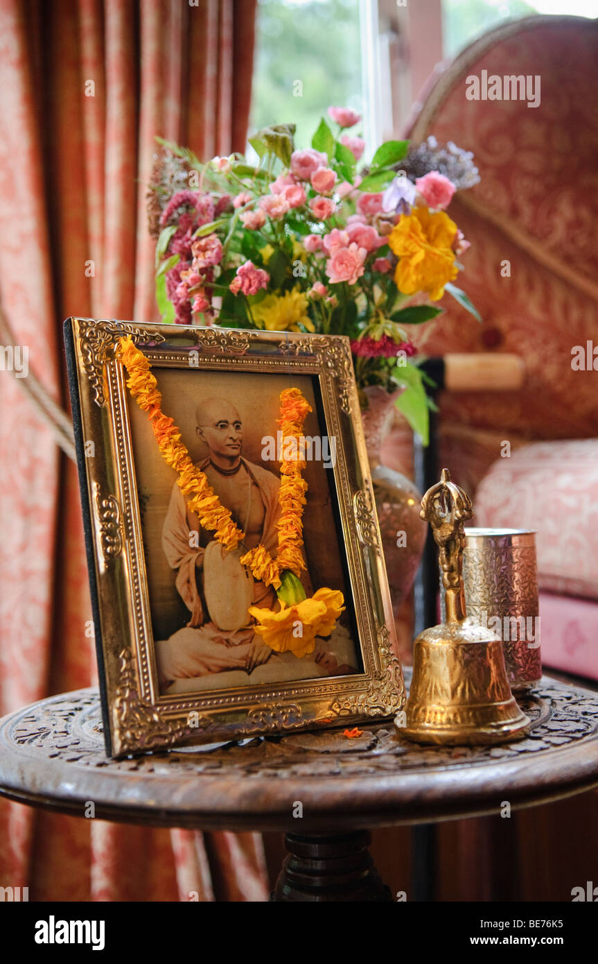 Photo d'un dévot Hare Krishna dans un cadre photo en laiton avec fleurs, une cloche et une tasse sur une table à un temple Banque D'Images